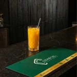 Coastal Coasters branded bar mat placed on a dark marble bar counter with a drink beside it.