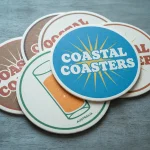 A stack of round branded drink coasters on a wooden surface, with the top blue coaster reading “Coastal Coasters” in white text over a yellow sunburst design.