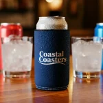 A chilled beverage can inside a navy blue Coastal Coasters stubby cooler on a wooden bar counter, with glasses and cans blurred in the background.