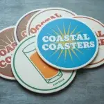 Stack of custom round coastal coasters featuring colorful branded designs, a sunburst logo, and an illustrated drink glass.