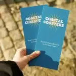 Hand holding two blue Coastal Coasters promotional brochures with white lettering and a gold sunburst design outdoors.