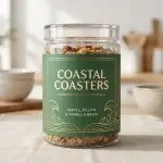 A clear jar of granola with a dark green and gold Coastal Coasters label reading “Premium Artisan Granola – Maple, Pecan & Vanilla Bean” on a kitchen counter.