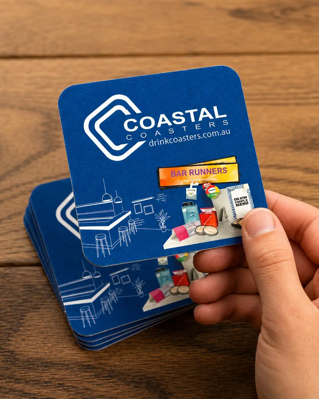 Hand holding a blue Coastal Coasters branded drink coaster above a stack of matching custom-printed coasters on a wooden table.